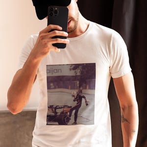 May include: A white t-shirt with a black and white photo of a race car driver on the front. The photo shows the driver in a racing suit, standing next to a race car. The text "aijan" is visible in the background.