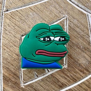 May include: A green frog meme with a sad expression, wearing a blue shirt. The frog has large, round eyes and a wide, open mouth.