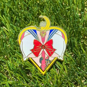 Op de afbeelding: Een gouden hartvormige pin met een wit en blauw matrozenuniform, een rode strik en een halve maan. De pin toont een cartoonfiguur in een roze en witte jurk met een zwarte kat.