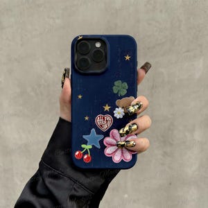 May include: A dark blue smartphone case featuring a variety of colorful stickers including a heart with the text ALL YOU NEED IS LOVE, a brown teddy bear, a green four-leaf clover, a white daisy, a blue star, a pink flower, and two red cherries.