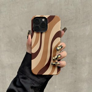 May include: A smartphone case featuring a wavy, abstract pattern in shades of dark brown, medium brown, and beige. The case has a cutout for a triple-lens camera system on the back, with a smooth, matte finish covering the entire surface area.