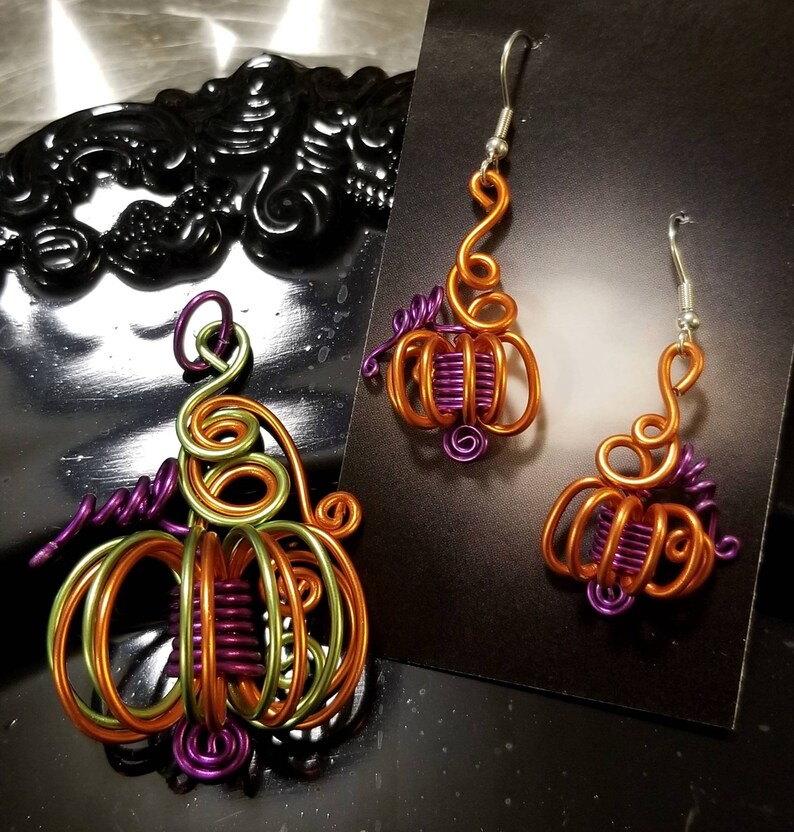 May include: A pair of pumpkin-shaped earrings and a pendant made of wire wrapped in orange, green, and purple wire. The earrings and pendant are handmade and have a whimsical design.