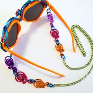 May include: Orange sunglasses with a colorful wire frame and a chain with colorful wire swirls. The chain is attached to the sunglasses with a metal loop.