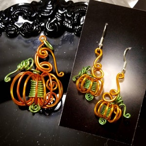 May include: A pair of pumpkin-shaped earrings and a matching pendant made of wire. The earrings and pendant are orange and green.