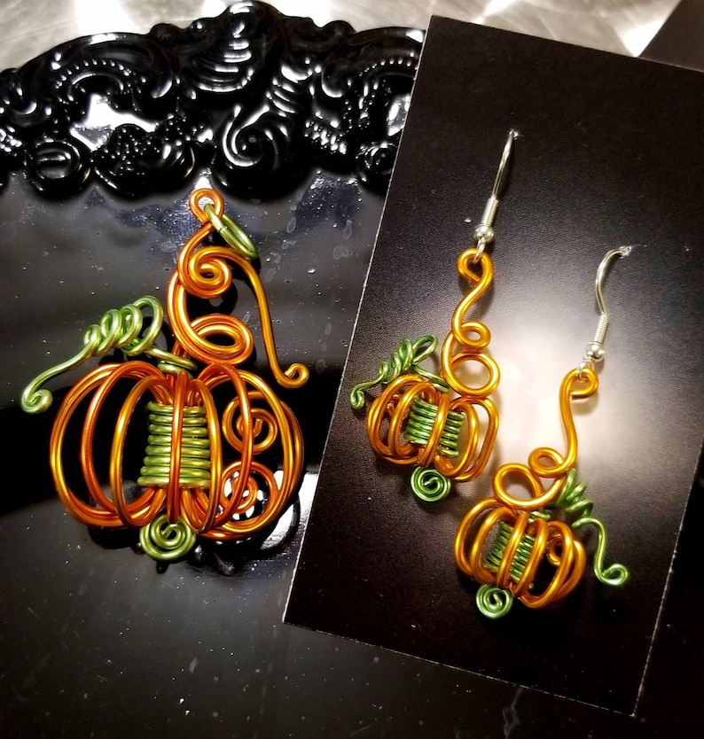 May include: A pair of pumpkin-shaped earrings and a matching pendant made of wire. The earrings and pendant are orange and green.