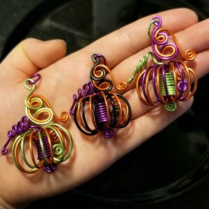 May include: Three wire-wrapped pumpkin ornaments in orange, green, and purple. Each pumpkin has a stem and is decorated with swirls of wire.