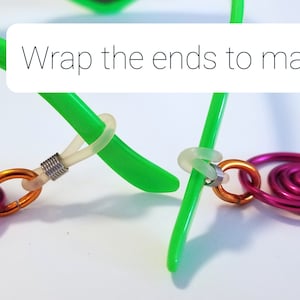 May include: Close-up of a green glasses frame with a pink and orange wire chain attached. The ends of the chain are wrapped around the glasses frame to make it tight. "Wrap the ends to make tight!" is written in white text on a white background.