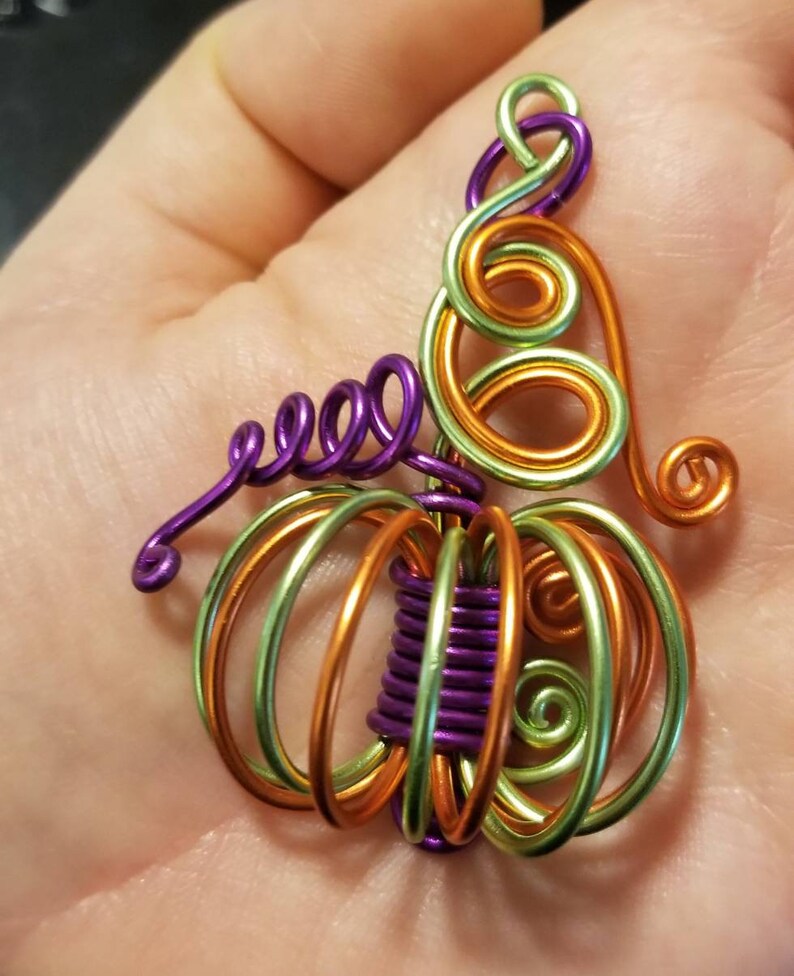 May include: A wire-wrapped pumpkin charm with green, orange, and purple wire. The charm is shaped like a pumpkin with a stem and leaves.