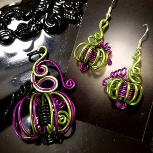 May include: A pair of green and purple wire pumpkin earrings. The earrings are made of wire that has been twisted and shaped into the form of a pumpkin. The earrings are hanging from silver hooks.