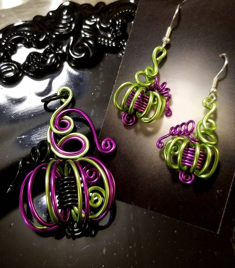 May include: A pair of green and purple wire pumpkin earrings. The earrings are made of wire that has been twisted and shaped into the form of a pumpkin. The earrings are hanging from silver hooks.