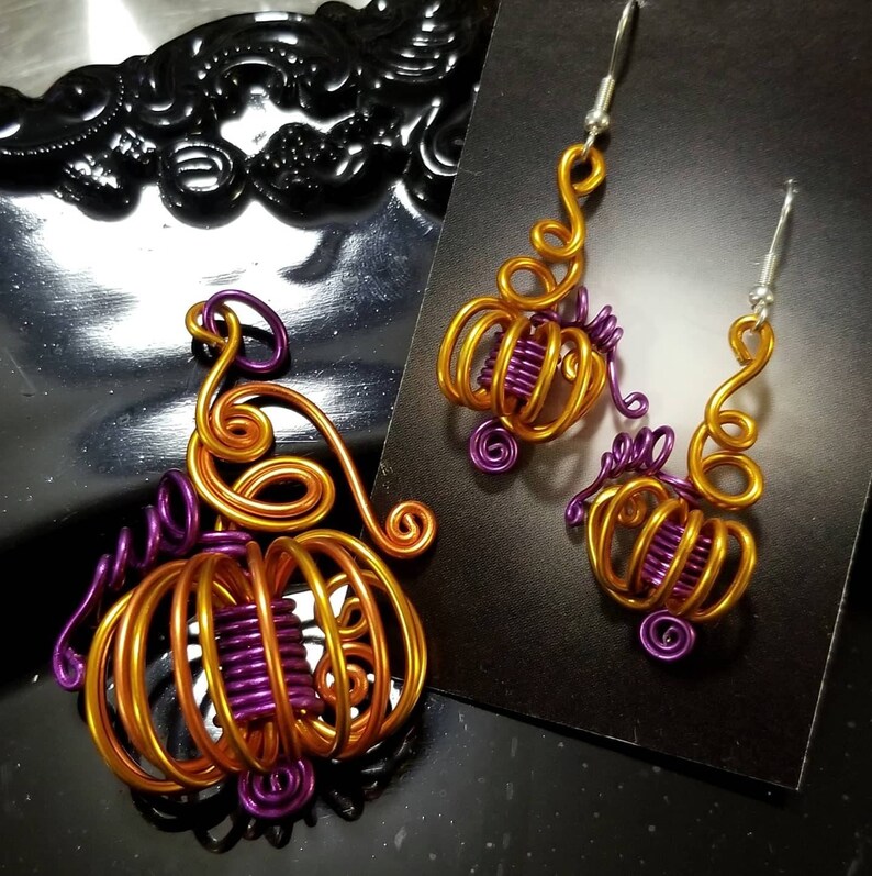 May include: A set of gold and purple wire pumpkin earrings and a matching pendant. The earrings are dangle style and the pendant is a charm.
