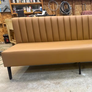 May include: A tan-colored, long bench with a vertically ribbed back and a smooth seat. The bench has black legs and is suitable for a dining area or waiting room. The bench measures approximately 72 inches long.