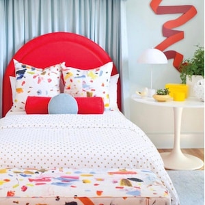 May include: A bedroom scene featuring a red headboard, patterned pillows, and a bedspread with gold dots. A bench with colorful abstract art sits at the foot of the bed. A side table holds a lamp and decorative items, with a ribbon-like wall art piece.