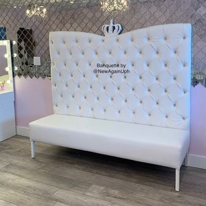 Op de afbeelding: Witte, gestoffeerde bank met een hoge, gebogen rugleuning. De rugleuning is versierd met een kroon en strass steentjes. De bank staat op een houten vloer. De tekst "Banquette by @NewAgainUph" staat op de rugleuning.