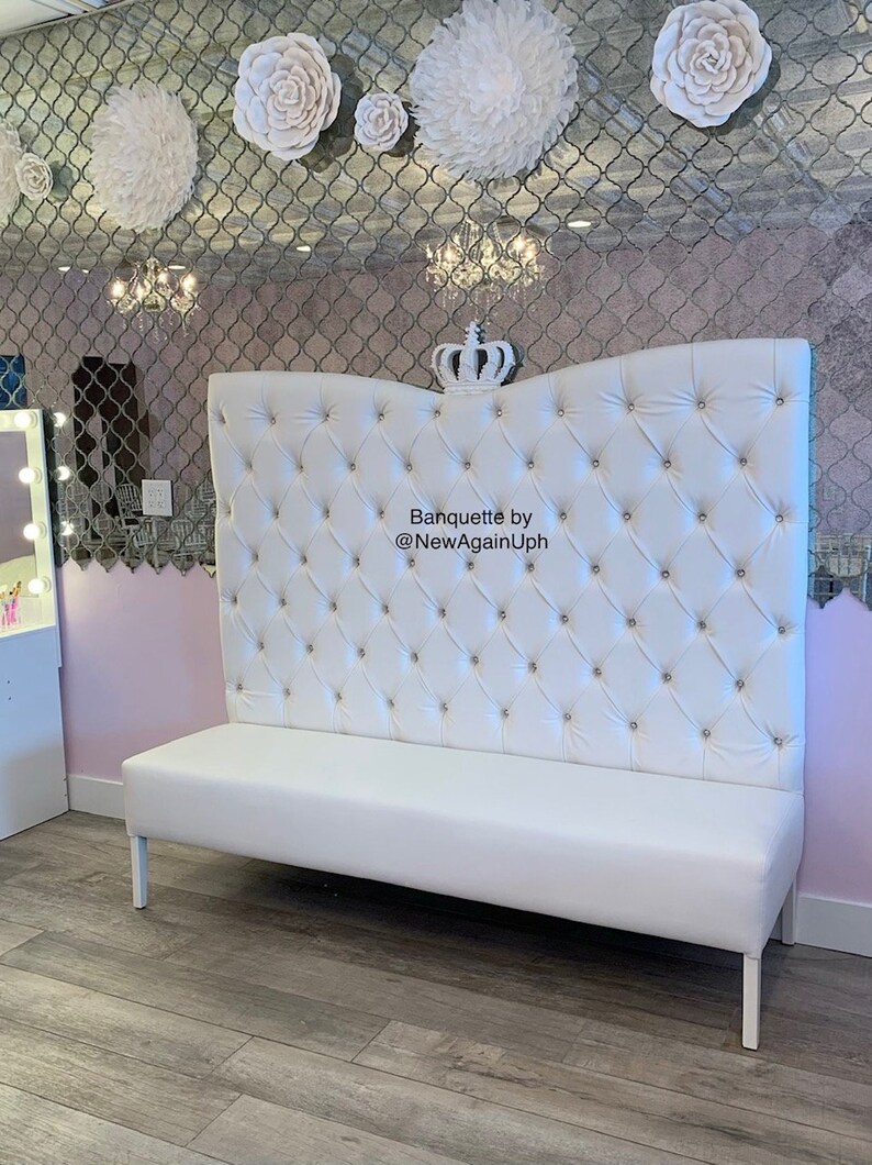 Op de afbeelding: Witte, gestoffeerde bank met een hoge, gebogen rugleuning. De rugleuning is versierd met een kroon en strass steentjes. De bank staat op een houten vloer. De tekst "Banquette by @NewAgainUph" staat op de rugleuning.