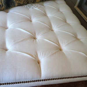 K&ouml;nnte beinhalten: Ein wei&szlig;er, gekn&ouml;pfter Ottoman mit braunem Rand. Der Ottoman hat ein Knopf-Steppdesign und ist aus einem weichen, pl&uuml;schigen Stoff gefertigt.