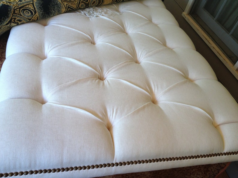 K&ouml;nnte beinhalten: Ein wei&szlig;er, gekn&ouml;pfter Ottoman mit braunem Rand. Der Ottoman hat ein Knopf-Steppdesign und ist aus einem weichen, pl&uuml;schigen Stoff gefertigt.
