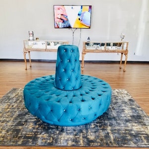 May include: A teal blue tufted round ottoman with a matching teal blue tufted top. The ottoman is on a gray and gold patterned rug.