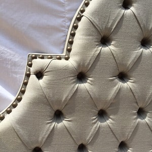 May include: A close-up of a tufted headboard with a diamond pattern and gold-colored nailhead trim. The headboard is made of a light beige fabric.