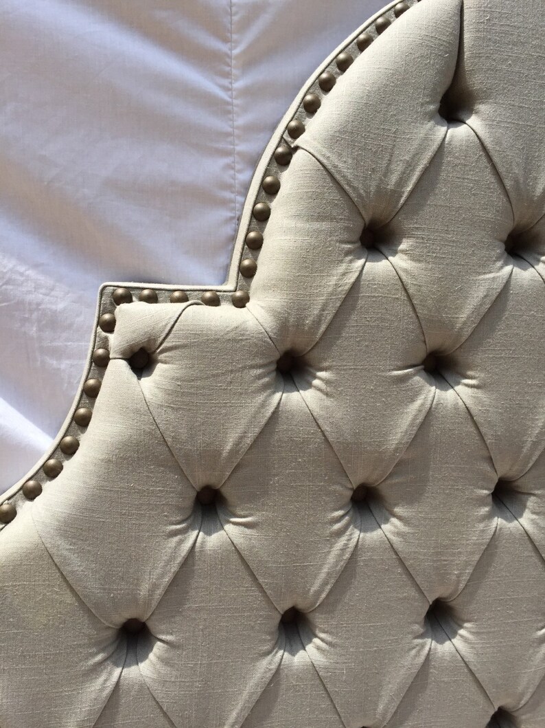 May include: A close-up of a tufted headboard with a diamond pattern and gold-colored nailhead trim. The headboard is made of a light beige fabric.