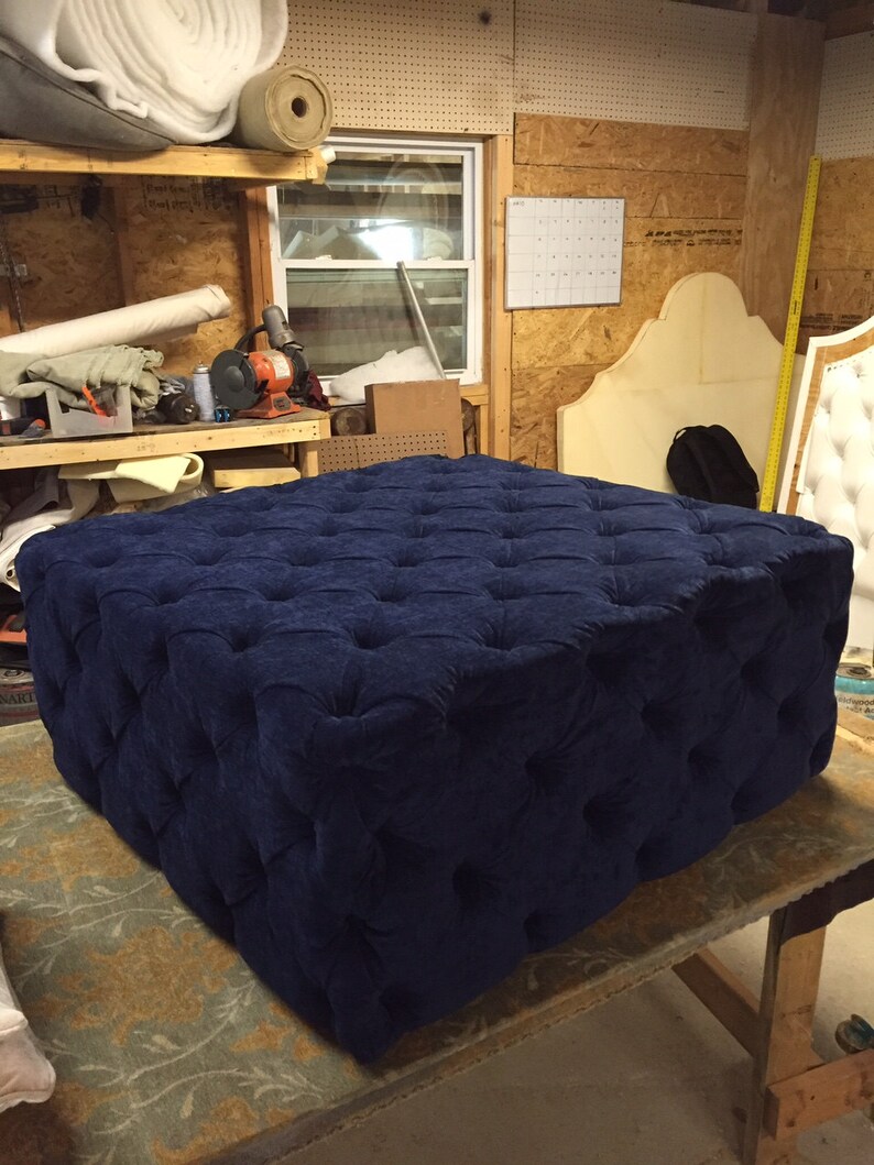 May include: A large, square, navy blue ottoman with tufted buttons. The ottoman is sitting on a wooden table with a patterned rug underneath.