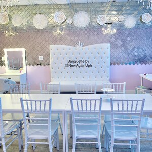 Op de afbeelding: Een witte bankettafel met witte stoelen, opgesteld in een kamer met een wit gestoffeerd hoofdeinde met een kroon erop. Het hoofdeinde heeft de tekst "Banquette by @NewAgainUph" erop. De kamer heeft een wit plafond met vierkante tegels en een gespiegelde muur met witte papieren bloemen.