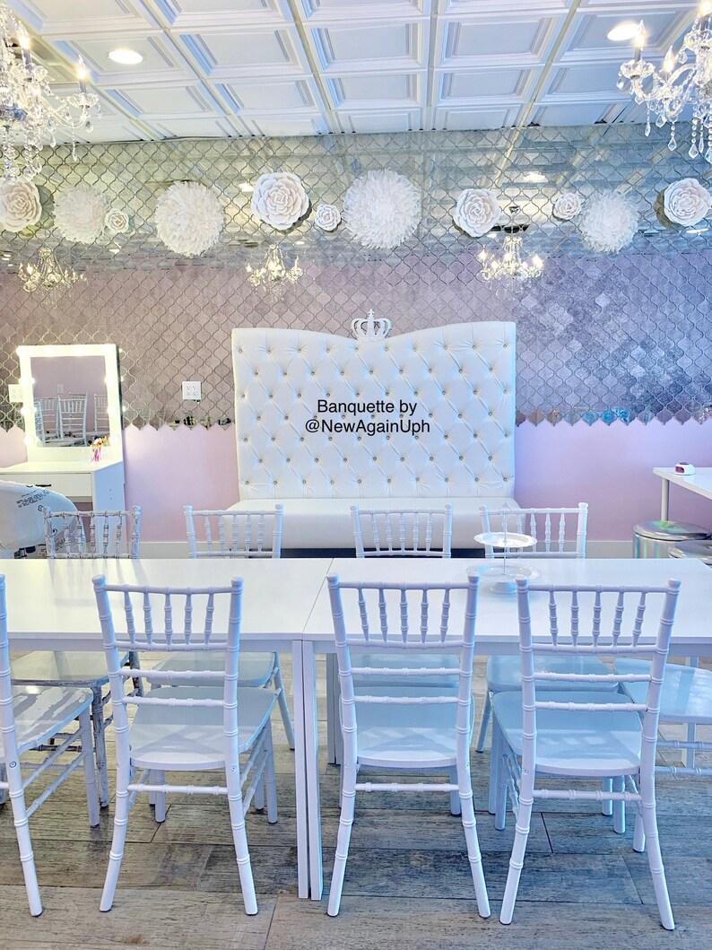 Op de afbeelding: Een witte bankettafel met witte stoelen, opgesteld in een kamer met een wit gestoffeerd hoofdeinde met een kroon erop. Het hoofdeinde heeft de tekst "Banquette by @NewAgainUph" erop. De kamer heeft een wit plafond met vierkante tegels en een gespiegelde muur met witte papieren bloemen.