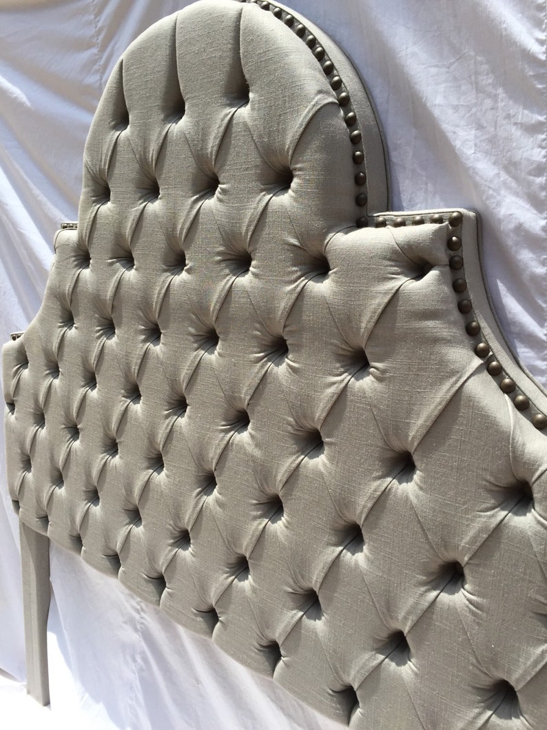 May include: A tufted headboard with a curved top and a linen fabric upholstery. The headboard is decorated with brass nailhead trim.
