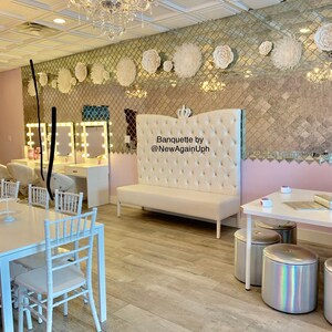 Op de afbeelding: Een witte, gestoffeerde bank met een kroon op de bovenkant, met het opschrift "Banquette by @NewAgainUph", in een kamer met witte stoelen en tafels, en een gespiegelde muur versierd met witte papieren bloemen.