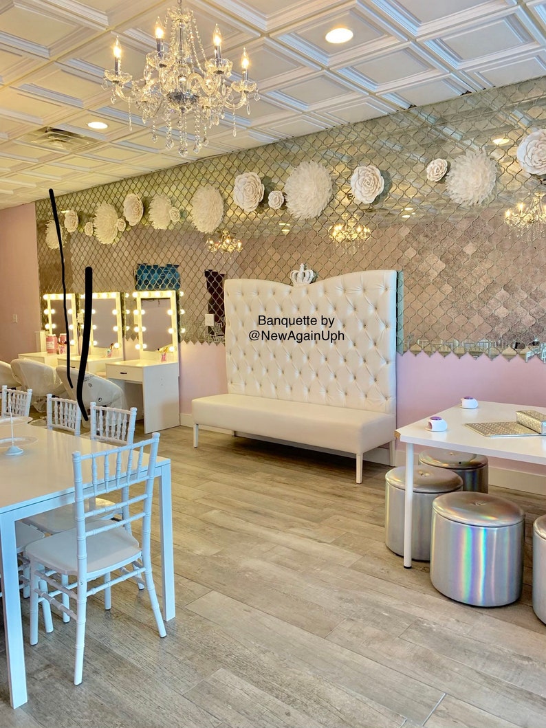 Op de afbeelding: Een witte, gestoffeerde bank met een kroon op de bovenkant, met het opschrift "Banquette by @NewAgainUph", in een kamer met witte stoelen en tafels, en een gespiegelde muur versierd met witte papieren bloemen.