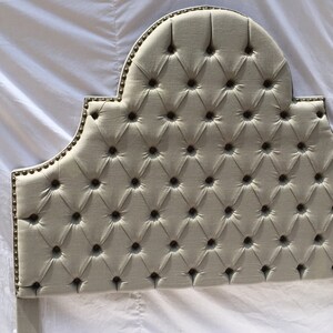 May include: A tufted headboard with a curved top and a diamond pattern. The headboard is upholstered in a light beige fabric and has silver-toned nailhead trim.