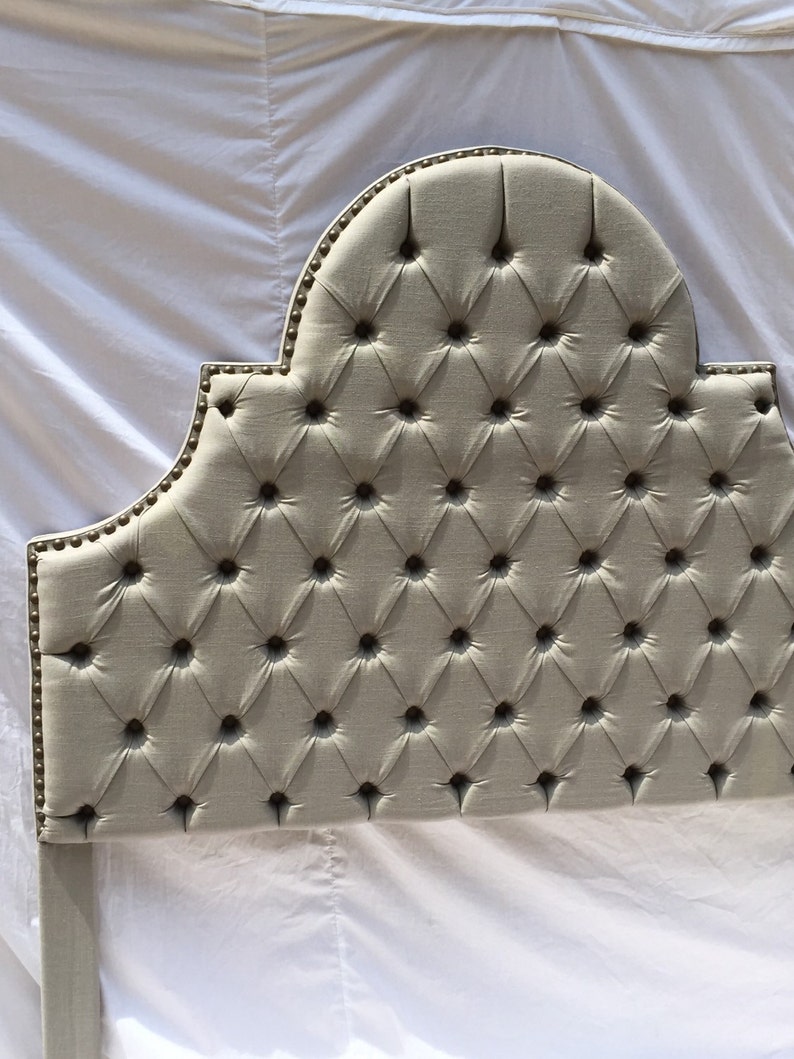 May include: A tufted headboard with a curved top and a diamond pattern. The headboard is upholstered in a light beige fabric and has silver-toned nailhead trim.