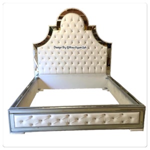 May include: A white upholstered bed frame with a mirrored headboard and footboard. The headboard features a tufted design and a mirrored arch. The footboard has a mirrored border and a tufted design. The text "Design By @NewAgainUph" is visible on the headboard.