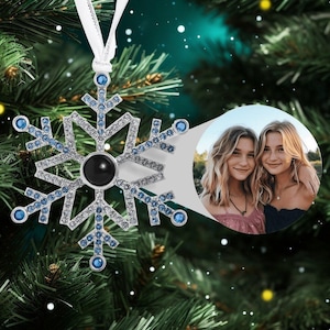 May include: A silver snowflake Christmas ornament with blue rhinestones and a black photo insert. The ornament has a white ribbon for hanging.