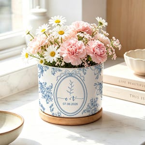 May include: A white ceramic planter with a blue floral pattern and a wooden base, filled with pink carnations, white daisies, and baby's breath. The planter features a central oval frame with the text C + T and the date 07.06.2026.