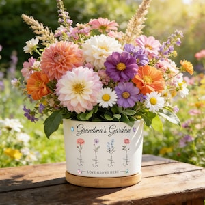 May include: A colourful floral display in a white ceramic pot, inscribed with "Grandma's Garden" and floral illustrations. The arrangement includes dahlias and daisies. The pot sits on a wooden surface, with a blurred garden background.