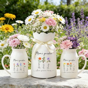 Puede incluir: Tres jarrones de cerámica blanca con arreglos florales. Los jarrones están inscritos con "Mom's garden" y "love grows here". El jarrón central tiene una parte superior festoneada y una cinta. Los otros dos jarrones tienen asas. Las flores incluyen margaritas, claveles y lavanda.