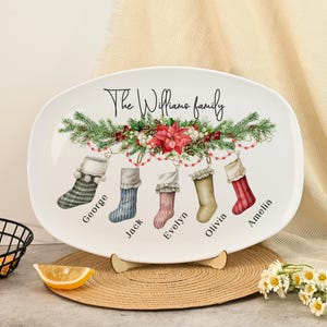 May include: Oval ceramic platter with a Christmas theme. The platter features a festive design with stockings, greenery, and the text "The Williams family." Each stocking is personalized with a name. The platter is supported by a wooden stand.