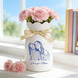 Puede incluir: Un jarrón de cerámica blanca con una parte superior festoneada, atado con una cinta dorada y con un boceto azul de una madre y un niño. El jarrón contiene claveles rosas y tiene el texto "I love you, Mom".