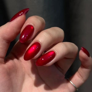 Puede incluir: Primer plano de una mano con uñas ovaladas rojas pintadas con un esmalte de uñas rojo oscuro brillante.