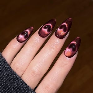 May include: A hand displaying long, almond-shaped nails in a rich burgundy hue. Each nail is adorned with a cat-eye effect, showcasing a heart-shaped design in pink and red tones, offering a distinctive and fashionable manicure.