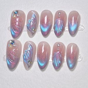 K&ouml;nnte beinhalten: Ein Set aus zehn k&uuml;nstlichen N&auml;geln mit rosa Basis und schillernden blauen und lila Akzenten. Jeder Nagel hat ein einzigartiges Design, einige mit Edelsteinverzierungen. Auf der Karte steht "Nail Art" und "Vielen Dank f&uuml;r Ihre Bestellung. Ich hoffe, es gef&auml;llt Ihnen."
