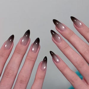 Puede incluir: Primer plano de manos con uñas largas en forma de estilete. Las uñas presentan un diseño degradado, pasando de un negro oscuro en las puntas a una base nude translúcida. Un anillo plateado con una pequeña piedra es visible en un dedo.