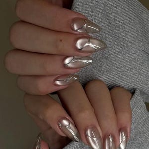 Puede incluir: Primer plano de manos con esmalte de uñas plateado metálico largo. Las uñas tienen forma de almendra con un acabado reflectante, similar al cromo. Finas líneas plateadas acentúan la superficie de las uñas. Las manos descansan sobre una tela gris.