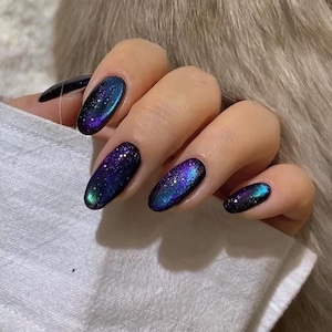Pode incluir: Unhas ovais com um design de galáxia. As unhas apresentam uma base azul escura com brilhantes detalhes roxos, verdes e prateados, criando um efeito cósmico. A mão está sobre um tecido branco.