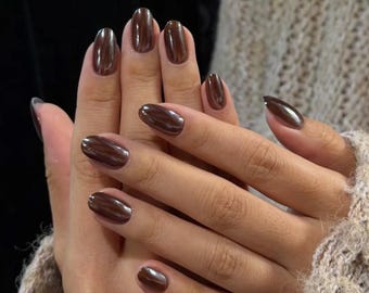 May include: Hands with polished, oval-shaped nails in a dark brown, almost metallic shade. The nails are glossy and reflect light, creating a sleek and modern look. The hands are resting, and the nails are the focal point.