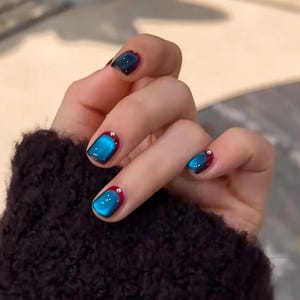 Könnte beinhalten: Eine Hand mit manikürten Nägeln. Die Nägel sind mit einem glänzenden Cat-Eye-Effekt in Blau- und Türkistönen lackiert, mit einer dunkelroten Basis und einem kleinen silbernen Punkt. Die Hand trägt einen dunkelbraunen Pullover.