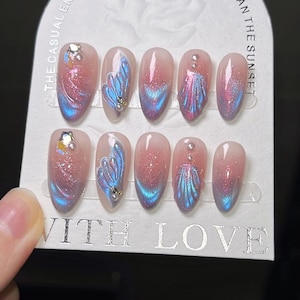 K&ouml;nnte beinhalten: Ein Set k&uuml;nstlicher N&auml;gel mit hellrosa Basis, verziert mit blau-rosa schillernden Designs, Glitzer und kleinen Verzierungen. Auf der Karte steht "The casual encounter is more gentle than the sunset" und "With Love".