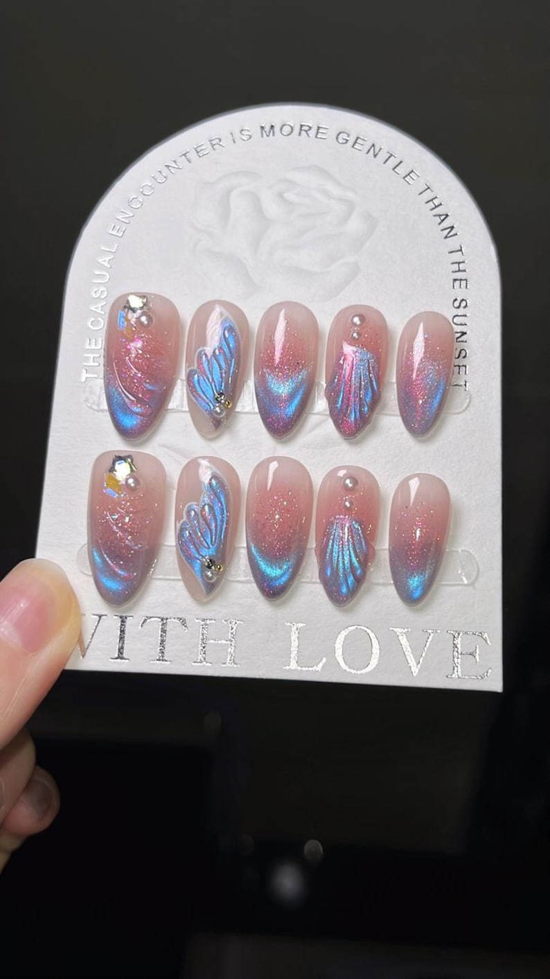 K&ouml;nnte beinhalten: Ein Set k&uuml;nstlicher N&auml;gel mit hellrosa Basis, verziert mit blau-rosa schillernden Designs, Glitzer und kleinen Verzierungen. Auf der Karte steht "The casual encounter is more gentle than the sunset" und "With Love".