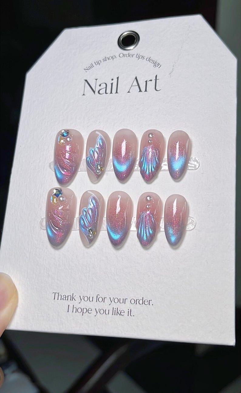 K&ouml;nnte beinhalten: Ein Set aus zehn k&uuml;nstlichen N&auml;geln mit rosa Basis und schillernden blauen und lila Akzenten. Jeder Nagel hat ein einzigartiges Design, einige mit Edelsteinverzierungen. Auf der Karte steht "Nail Art" und "Vielen Dank f&uuml;r Ihre Bestellung. Ich hoffe, es gef&auml;llt Ihnen."